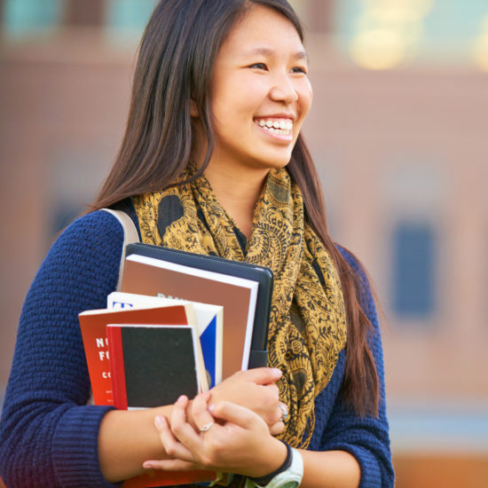 Shot of a young female student standing with her books on campus smiling and looking on; campus booksstore