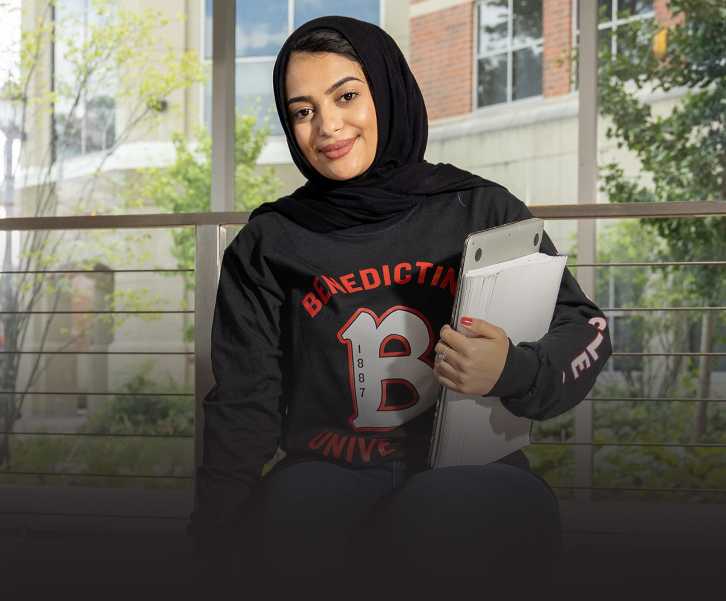 Female student sitting in campus building holding laptop and stack of papers, admitted, transfer, and degree completion programs