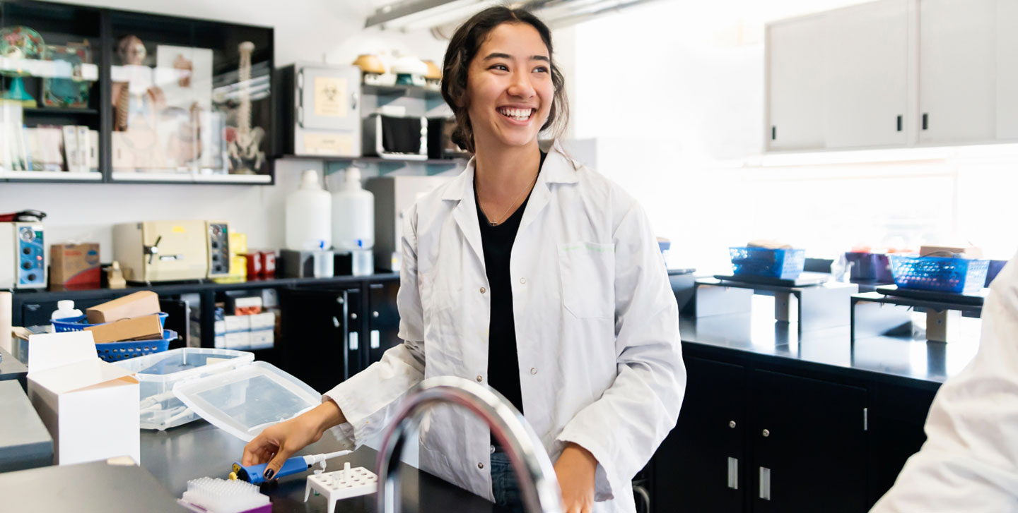 female student in lab coat smiling in lab reaching for a pipet; biology program