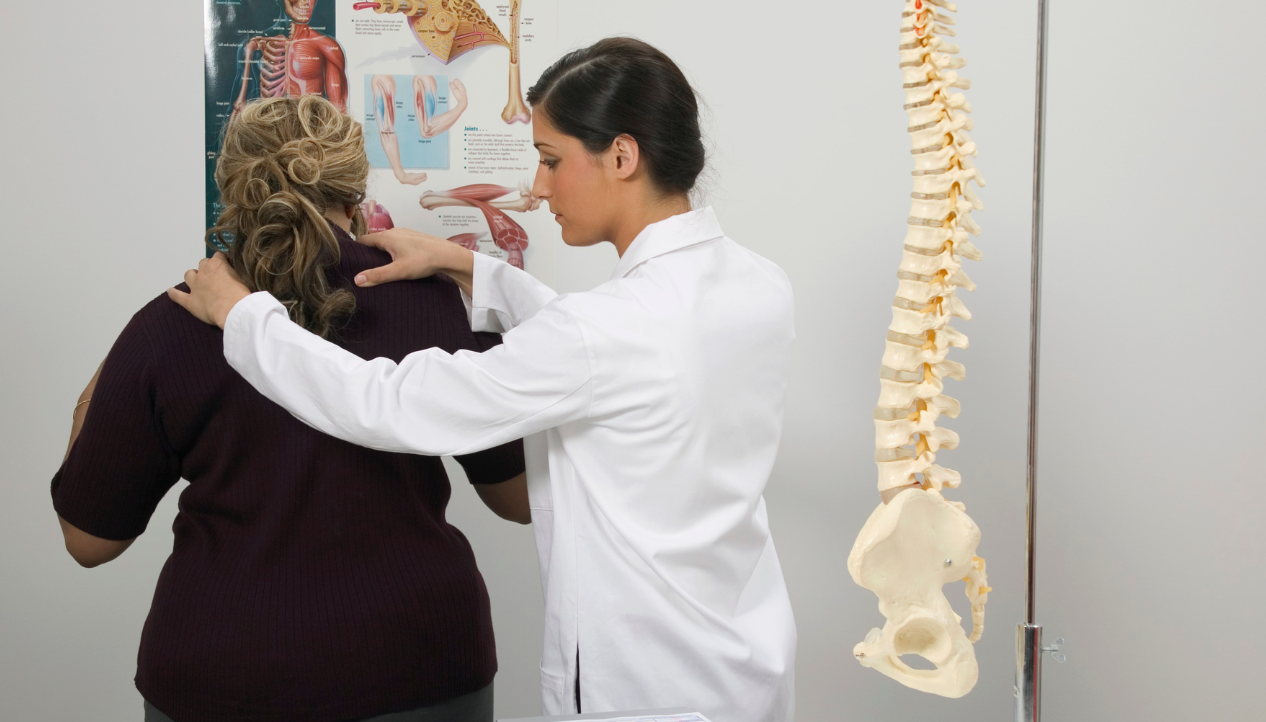 female chiropractor treating shoulders of patient, spine model hanging next to her; pre-chiropractic program