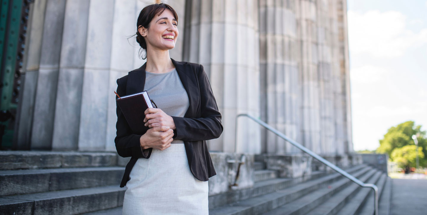 female professional holding book walking up steps of large building; criminology program