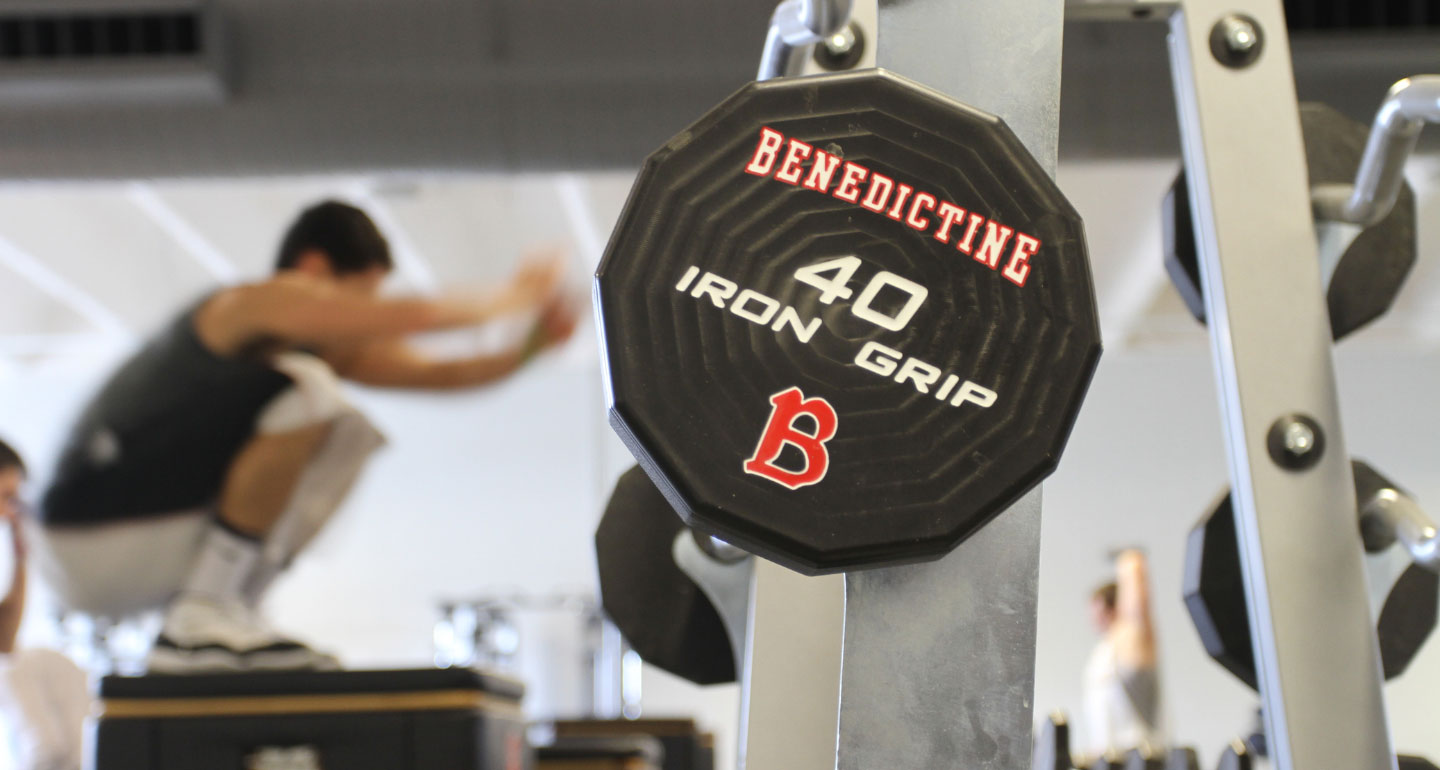 close up image of weight in Benedictine's weight room with student doing standing jump in the background; strength and conditioning minor