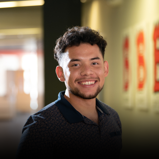 Head shot of Hispanic male student smiling in a hallway with Benedictine "B" images on the wall next to him; Transfer Degree scholarships