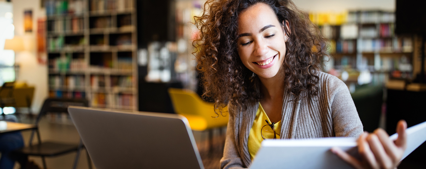 female student smiling looking at notebook sitting at laptop in the library