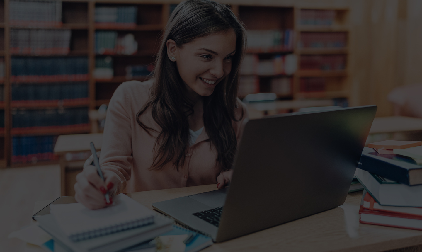 female student taking notes at her laptop; image has a dark gray overlay on it; library