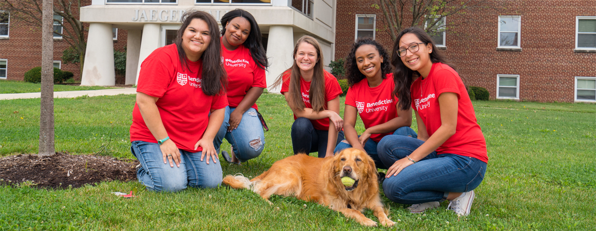 Group of five female students gathered around a dog with a ball on the quad; Res Life