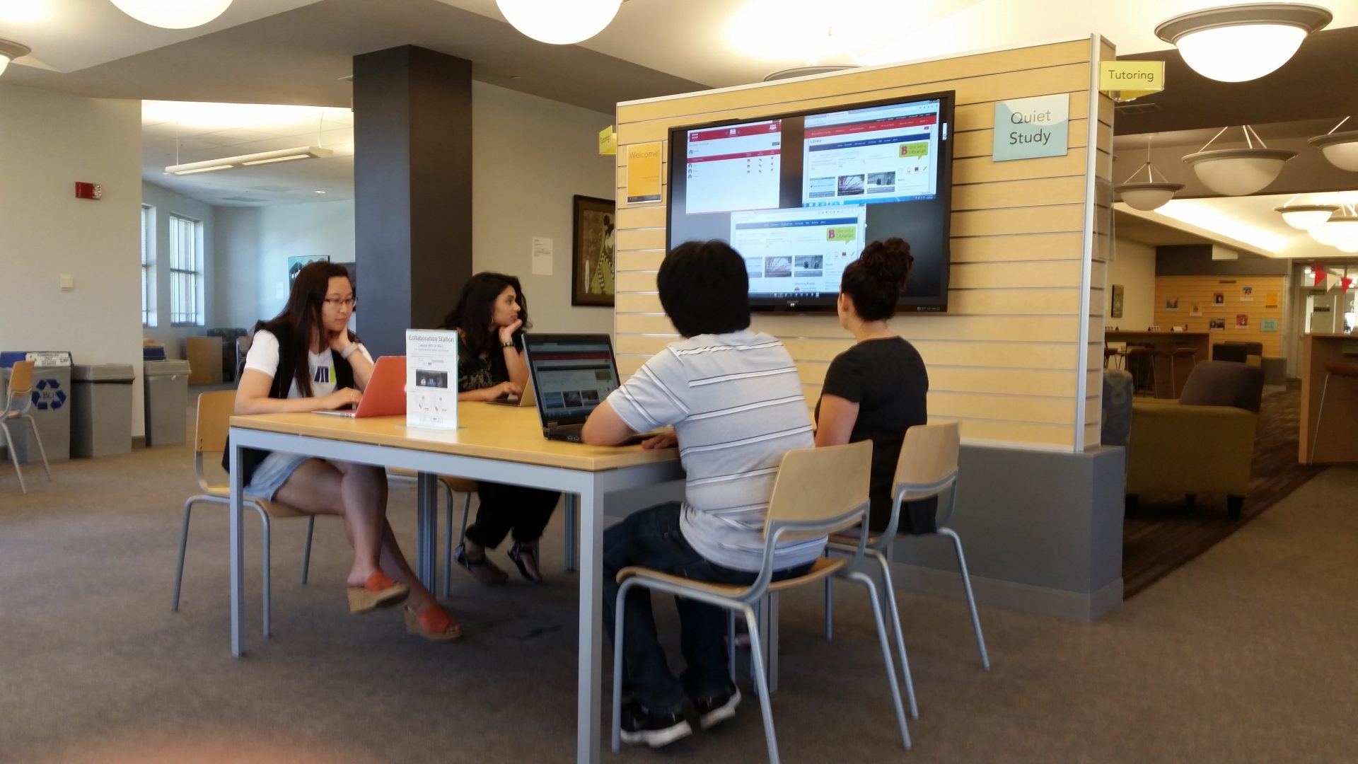 group of students sitting together at a table watching a large tv screen in the library