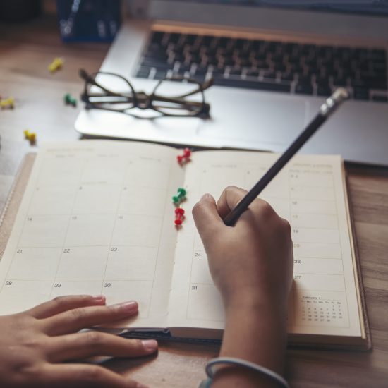 Hands writing in a calendar with a pencil, thumbtacks are scattered on the calendar and table - working from home