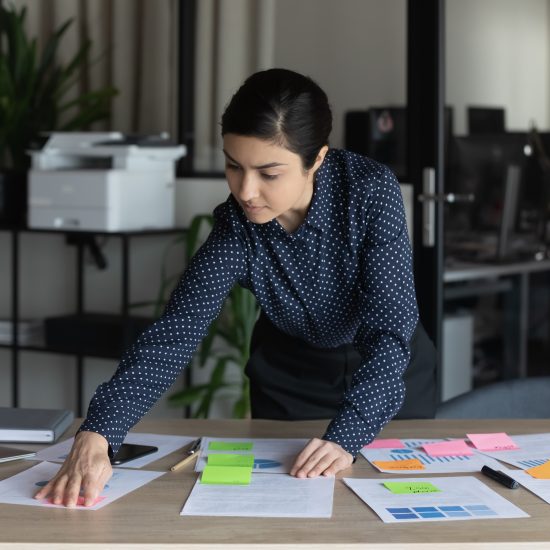 Confident Indian businesswoman working with project documents, colorful stickers, standing in office, focused employee executive analyzing financial statistics, reading notes, developing strategy