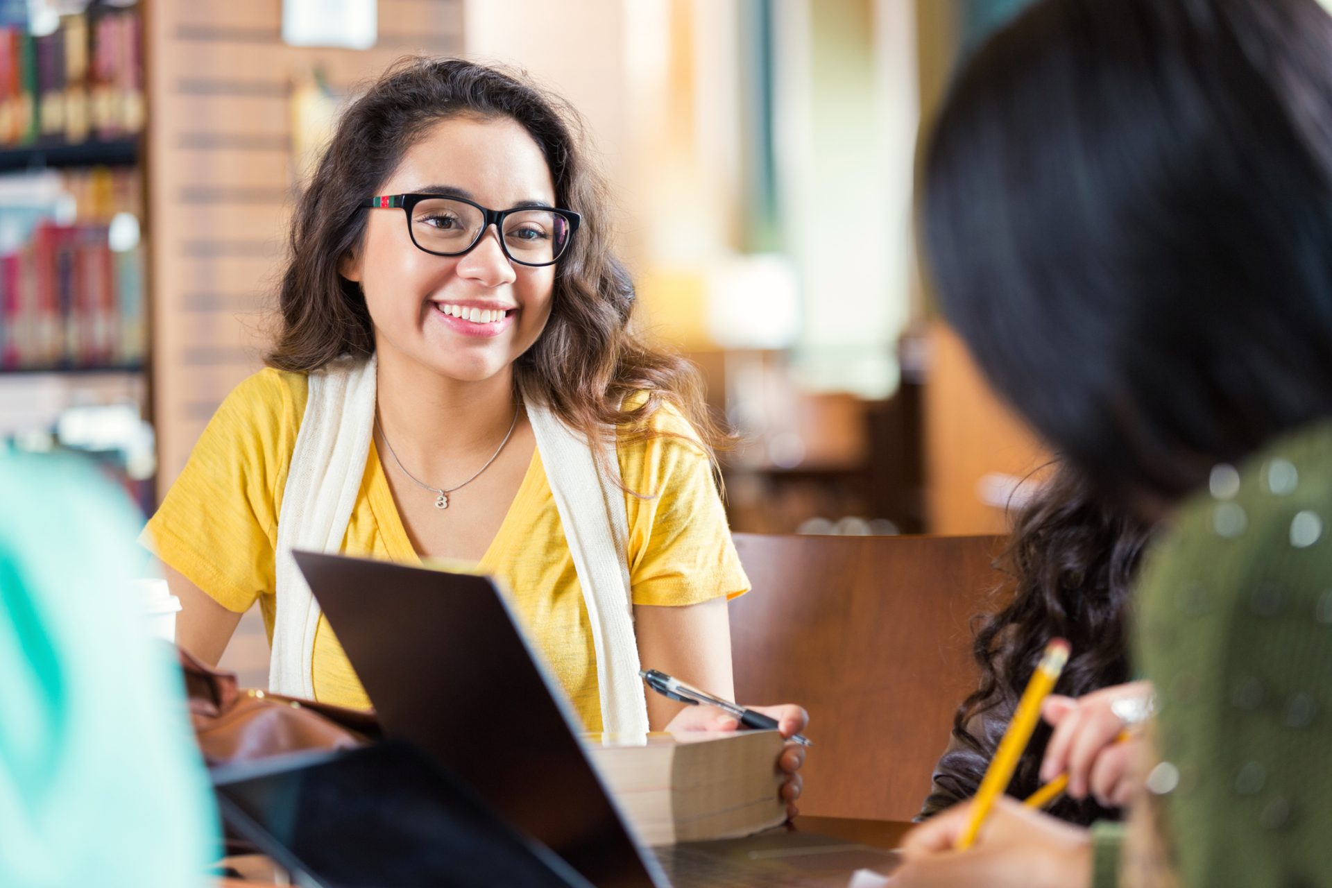 Trendy hipster Hispanic college age girl is smiling while sitting at tabe in library with group of friends. Students are working in study group to prepare for exam while using laptop computers, digital tablets, and other technology.