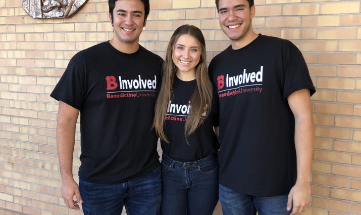 Three students with "B Involved" t-shirts posing in front of Mary wall plaque: Campus Ministry Mesa
