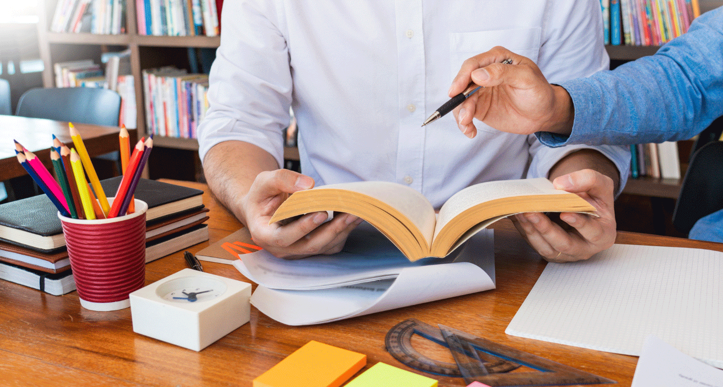 man sitting at desk with open book and another person with pen pointing at open book