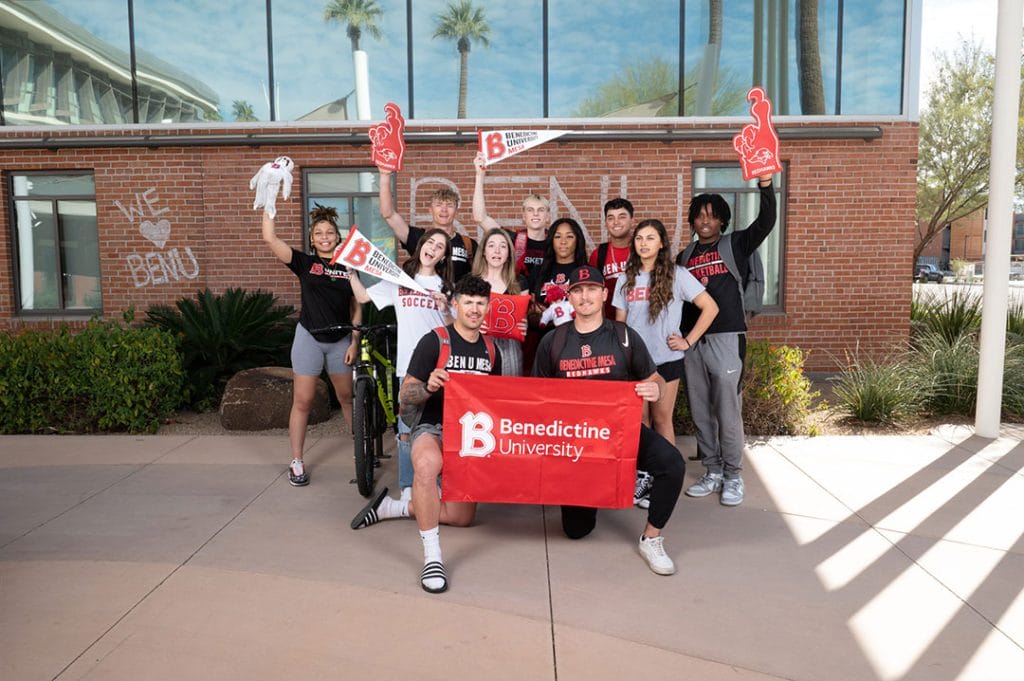 Mesa students with BenU spirit gear posing outside Gillett Building holding Benedictine banner